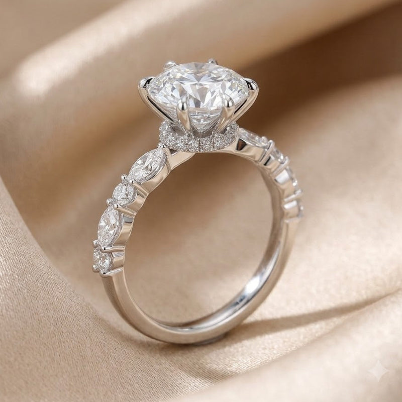 A custom platinum solitaire engagement ring with a large round-2.5ct brilliant cut natural diamond and marquis diamond accents along the band, displayed on draped champagne satin fabric. Firefly Jewelers design.